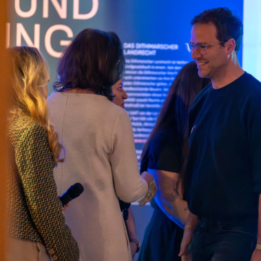 Vier Personen stehen in einem Raum und unterhalten sich. Eine Frau hält ein Mikrofon. Im Hintergrund ist ein beleuchtetes Schild mit teilweise lesbarem Text. Die Stimmung ist freundlich und die Beleuchtung warm.