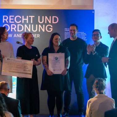 Eine Gruppe von sieben Personen steht auf einer Bühne. Zwei Frauen halten einen großen Scheck und eine Urkunde. Im Hintergrund ist ein Schild mit der Aufschrift "Recht und Ordnung" erkennbar. Die Szene wirkt festlich beleuchtet.