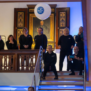Eine Gruppe von Menschen in schwarzen Outfits steht auf einer Treppe und einem Balkon in einem Gebäude mit Holzelementen. Im Hintergrund hängt ein Porträt. Ein Ballon mit einem Logo schwebt über ihnen.