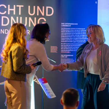 Drei Personen stehen vor einer blau beleuchteten Wand mit Aufschriften. Zwei Frauen geben sich die Hand, während die dritte Person daneben ein Mikrofon hält. Im Vordergrund sind schemenhaft zwei Köpfe zu erkennen.