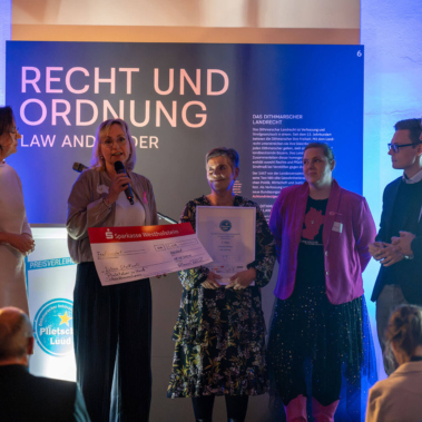 Eine Gruppe von Personen steht auf einer Bühne vor einem Publikum. Eine Person hält ein großes Scheck-Poster, eine andere eine Urkunde. Im Hintergrund ist ein Banner mit der Aufschrift „Recht und Ordnung“.