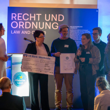 Eine Gruppe von Personen steht auf einer Bühne bei einer Preisverleihung. Eine Frau hält ein Mikrofon und spricht. Ein großer Scheck wird hochgehalten. Im Hintergrund befinden sich ein Rednerpult und ein beleuchtetes Poster mit "Recht und Ordnung".