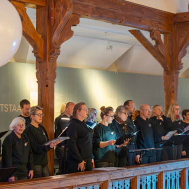Eine Gruppe von Menschen steht in einem Raum mit holzverzierten Balken. Sie tragen dunkle Kleidung und halten Notenblätter in den Händen, während sie singen oder sprechen. Im Hintergrund ist eine grüne Wand zu sehen.