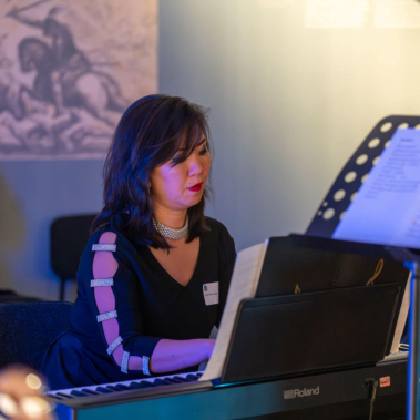 Eine Person spielt auf einem E-Piano und liest von einem Notenständer. Sie trägt ein schwarzes Kleid und eine Halskette. Im Hintergrund sitzt eine weitere Person, und an der Wand hängt ein gerahmtes Bild. Mikrofon- und Bühnenbeleuchtung sind sichtbar.