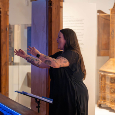 Eine Frau mit langen Haaren und Tätowierungen steht in einem Raum mit Holzelementen. Sie gestikuliert mit den Händen nach vorne. Im Hintergrund sind Vitrinen und ein Möbelstück sichtbar. Die Szene ist in warmem Licht beleuchtet.