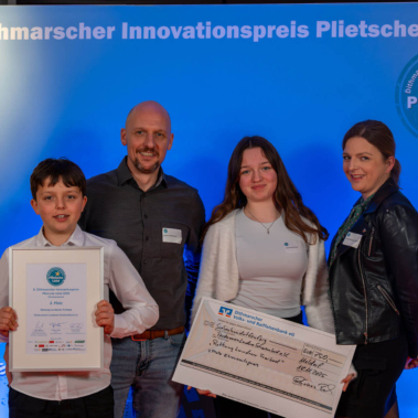 Eine Gruppe von vier Personen steht lächelnd nebeneinander vor einem blauen Hintergrund mit der Aufschrift "Dithmarscher Innovationspreis Plietsche Lüüd". Eine Person hält eine Urkunde, eine andere einen großen Scheck.
