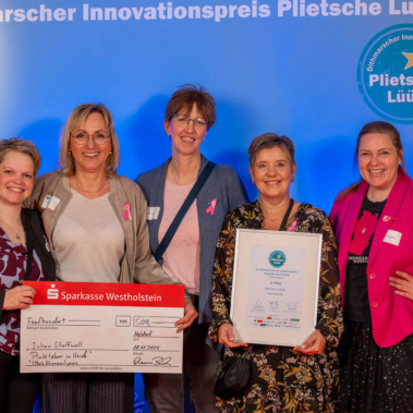 Fünf Personen lächeln in die Kamera. Eine hält einen großen Scheck und eine andere ein gerahmtes Zertifikat. Im Hintergrund ist ein Banner mit dem Logo des Dithmarscher Innovationspreises "Plietsche Lüüd" zu sehen.