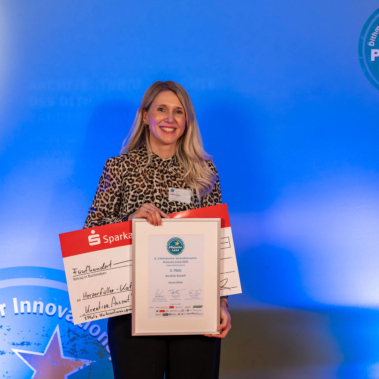 Eine lächelnde Frau hält eine gerahmte Urkunde und einen großen roten Scheck in den Händen. Der Hintergrund ist blau mit einem runden Logo auf der rechten Seite. Sie trägt eine gemusterte Bluse.