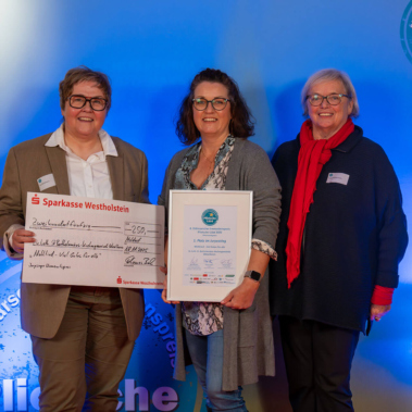 Drei Personen stehen nebeneinander vor einem blauen Hintergrund. Eine Person hält einen großen symbolischen Scheck, eine andere eine gerahmte Urkunde. Alle lächeln in die Kamera.