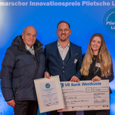 Drei Personen stehen vor einem blauen Hintergrund mit der Aufschrift "Dithmarscher Innovationspreis". Eine Person hält eine Urkunde, eine andere einen großen Scheck. Alle lächeln in die Kamera.