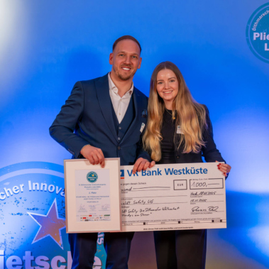 Ein Mann und eine Frau stehen lächelnd nebeneinander. Beide halten ein Zertifikat und einen großen symbolischen Scheck. Im Hintergrund ist eine blaue Wand mit dem Logo "Plietsche Lüü" und Regale mit Büchern.