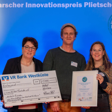 Drei Personen stehen vor einem blauen Hintergrund mit einem Schild "Dithmarscher Innovationspreis Plietsche Lüüd". Eine Person hält einen großen Scheck, eine andere eine gerahmte Urkunde. Alle lächeln in die Kamera.