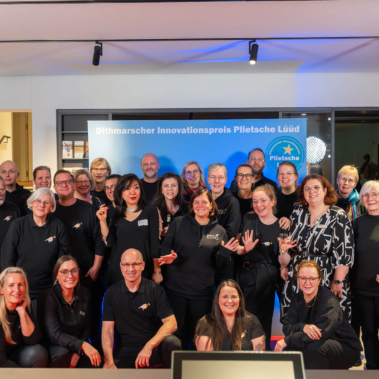 Eine Gruppe von Männern und Frauen in schwarzen Outfits lächelt in die Kamera. Sie stehen vor einem bunten Hintergrund mit der Aufschrift "Dithmarscher Innovationspreis Pilotsche Lüüd".