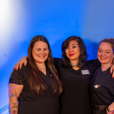 Drei lächelnde Personen in schwarzer Kleidung stehen nebeneinander und legen die Arme um die Schultern der anderen. Der Hintergrund ist blau beleuchtet. Links im Bild sind Bücherregale mit bunten Büchern zu sehen.