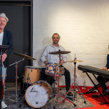 Drei Männer musizieren zusammen: Einer spielt Kontrabass, ein zweiter sitzt am Schlagzeug und ein dritter spielt Klavier. Sie befinden sich auf einem roten Teppich vor einer weißen Backsteinwand.