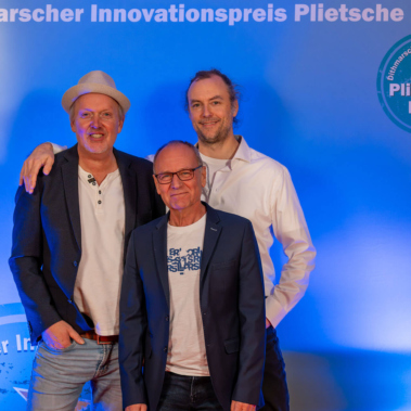 Drei Männer stehen vor einer blauen Hintergrundwand mit der Aufschrift "Dithmarscher Innovationspreis Plietsche Lüüd". Der Mann links trägt einen Hut, der Mann rechts hat die Hand auf der Schulter des Mannes in der Mitte.