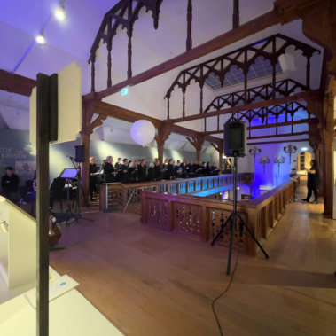 Eine Gruppe von Menschen steht in einem Raum mit Holzarchitektur und bemalter Decke. Im Vordergrund ist ein offener Bereich mit Geländer und blauem Licht. Links steht ein Stativ mit Mikrofon, und im Hintergrund sind thematische Ausstellungsstücke sichtbar.