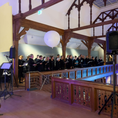 Eine Gruppe von Menschen steht auf einem hölzernen Balkon und singt in einem offenen Raum mit gotischen Stilelementen. Im Vordergrund ist ein Mikrofonstativ. Oben hängt ein großer, weißer Ballon.